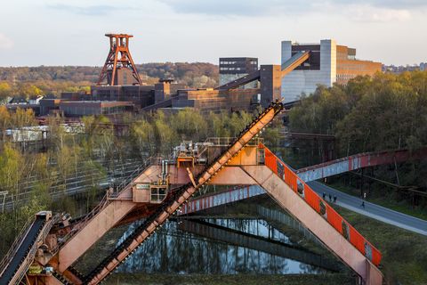 Die Geschichte Zollvereins beginnt in den Pioniertagen des heutigen Ruhrgebiets. Der erste Schacht wurde 1847 abgeteuft, vier Jahre später die erste Kohle gefördert: Vier weitere Anlagen entstanden, die letzte war die Schachtanlage Zollverein XII,  die ab 1932 die Förderung der gesamten Zollverein-Kohle zentral übernahm. 

Die Gebäude der 1928 bis 1932 im Stil der „Neuen Sachlichkeit“ nach Entwürfen der Architekten Fritz Schupp und Martin Kremmer gebauten Anlage sind nach dem Prinzip „Form follows Function“ zum optimalen Ablauf der Kohleförderung und -aufbereitung angeordnet. Trotz Rekordzahlen wie 12.000 Tagestonnen verwertbarer Steinkohle machte das Ende der Schwerindustrie auch vor Zollverein nicht Halt. Am 23. Dezember 1986 fuhr die letzte Schicht ein. An diesem Tag stand die Zeche bereits unter Denkmalschutz, 1990 begannen die Sanierungsarbeiten und seit 2001 sind Zeche und Kokerei Zollverein UNESCO-Welterbe. Mit dem Denkmalpfad ZOLLVEREIN®, dem Ruhr Museum und dem Red Dot Design Museum sind auf Schacht XII drei der beliebtesten Touristenziele der Region angesiedelt. Das Doppelbock-Fördergerüst der Schachtanlage ist heute ein Wahrzeichen für das gesamte Ruhrgebiet. 

Der Portalkratzer zwischen dem Zechengelände von Schacht XII und der Kokerei wurde 1973 auf dem Kohlenlagerplatz installiert. Es handelt sich um ein sogenanntes Kombinationsgerät der Firma Schade, das gleichzeitig einen Absetzer zum Haldenaufbau sowie einen Kratzer zum Abbau der Halde beinhaltet. Der Absetzer hat die von Zollverein Schacht XII kommende Kokskohle zu einer Halde aufgebaut, während der Kratzer mit an Ketten befestigten Schaufeln die Halde abgekratzt und auf ein laufendes Förderband weiter zur Kokerei transportiert hat. Die Kokskohle wurde gelagert, um im Falle eines Produktionsstillstandes auf der Zeche weiterhin die Kokerei mit Kokskohle zu versorgen, denn die Koksöfen mussten permanent produzieren und durften nicht erkalten.

Ort: Areal A [Schacht XII]