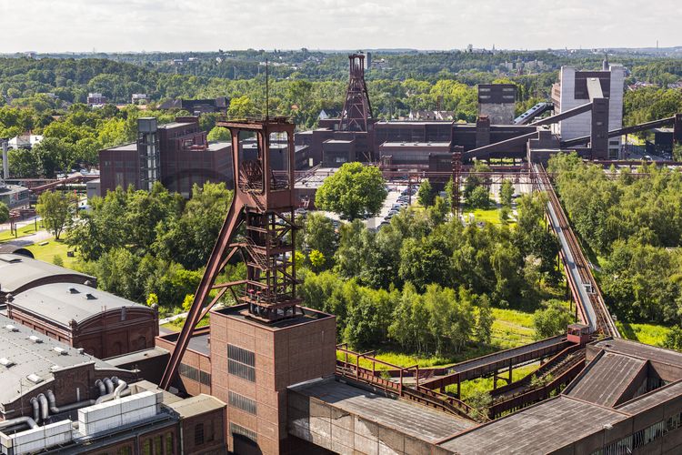 Zollverein Schacht XII
Die Geschichte Zollvereins beginnt in den Pioniertagen des heutigen Ruhrgebiets. Der erste Schacht wurde 1847 abgeteuft, vier Jahre später die erste Kohle gefördert: Vier weitere Anlagen entstanden, die letzte war die Schachtanlage Zollverein XII,  die ab 1932 die Förderung der gesamten Zollverein-Kohle zentral übernahm. 

Die Gebäude der 1928 bis 1932 im Stil der „Neuen Sachlichkeit“ nach Entwürfen der Architekten Fritz Schupp und Martin Kremmer gebauten Anlage sind nach dem Prinzip „Form follows Function“ zum optimalen Ablauf der Kohleförderung und -aufbereitung angeordnet. Trotz Rekordzahlen wie 12.000 Tagestonnen verwertbarer Steinkohle machte das Ende der Schwerindustrie auch vor Zollverein nicht Halt. Am 23. Dezember 1986 fuhr die letzte Schicht ein. An diesem Tag stand die Zeche bereits unter Denkmalschutz, 1990 begannen die Sanierungsarbeiten und seit 2001 sind Zeche und Kokerei Zollverein UNESCO-Welterbe. Mit dem Denkmalpfad ZOLLVEREIN®, dem Ruhr Museum und dem Red Dot Design Museum sind auf Schacht XII drei der beliebtesten Touristenziele der Region angesiedelt. Das Doppelbock-Fördergerüst der Schachtanlage ist heute ein Wahrzeichen für das gesamte Ruhrgebiet. 

Die 1851 in Betrieb genommene Gründerschachtanlage 1/2/8 ist die älteste Schachtanlage der Zeche Zollverein. Die vorhandenen Ziegelmassivbauten stammen aus dem ersten Jahrzehnt des 20. Jahrhunderts, sämtliche Erweiterungsbauten nach 1932 wurden im architektonischen Stil der Schachtanlage Zollverein XII errichtet. Auf Schacht 1/2/8 befanden sich mit der Markenkontrolle, Kaue und Lampenstube auch die Sozialräume für die auf Schacht 1/2/8 und Schacht XII tätigen Bergleute. Nach der Stilllegung 1986 wurde die Schachtanlage 1/2/8 saniert und beherbergt heute Ateliers sowie die renommierte Kulturinstitution PACT Zollverein. 2000 wurde die Schachtanlage 1/2/8 in die Denkmalliste der Stadt Essen aufgenommen und gehört seit 2001 zum UNESCO-Welterbe. 

Ort: Areal B [Schacht 1/2/8]