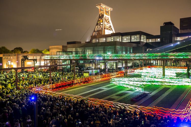 Die Performance „Speed of Light“ der schottischen Künstlerorganisation Nationale Vitae Activa (NVA) ließ am 4. Oktober 2013 mit 120 Läufer in LED-Lichtanzügen das UNESCO-Welterbe Zollverein in ganz neuem Licht erstrahlen.

Gleisboulevard nennt sich der Platz nördlich der Kohlenwäsche zwischen der Fritz-Schupp-Allee und der Halle 12 [A12]. Zu Betriebszeiten reihten sich dort die Waggons aneinander, die unter den Produktionsanlagen mit Kohle bzw. Bergematerial beladen wurden. 

Die Mannschaftsbrücke führte von der Schachtanlage 1/2/8 zur Schachtanlage XII und war der Verbindungsweg für die Bergleute. Ein Passieren zu ebener Erde war aufgrund des Zugverkehrs nicht möglich.

Pause machen im Bistro „Butterzeit!“ und Kunsthandwerk entdecken, wo früher Waggons mit Kohle beladen wurden: Die Ateliers im Erdgeschoss der Halle 12 [A12], darunter SchmuckProdukt und die ARKA Kulturwerkstatt, präsentieren neben eigenen Arbeiten auch wechselnde Ausstellungen anderer Künstler. Im Obergeschoss der ehemaligen Lesebandhalle finden heute Kongresse, Messen und Konzerte statt. Bis in die 1960er Jahre lasen hier Bergleute an fünf Lesebändern von Hand die großen Gesteinsstücke aus dem Rohkohlegemisch.

Das 55 Meter hohe Doppelbock-Fördergerüst ist das Wahrzeichen der Zeche Zollverein, der Stadt Essen und der gesamten Region: Die als Doppelstrebengerüst in Vollwandbauweise konstruierte Förderanlage ermöglichte eine doppelte Fördermechanik, mittels derer die doppelte Menge Förderwagen bzw. Fördergefäße transportiert werden konnten. Zollverein Schacht XII galt zu Betriebszeiten als die leistungsstärkste Zeche der Welt. 250 Stufen führen auf die oberste Plattform des Gerüstes, deren Seilscheiben sich auch heute noch drehen: Der Schacht wird heute von der RAG AG für die zentrale Wasserhaltung genutzt.

Ort: Areal A [Schacht XII], Gleisboulevard