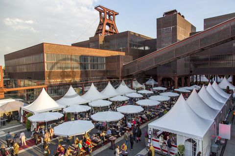 Einmal im Jahr treffen die regionalen Spitzengastronomen zur Gourmet-Meile Metropole Ruhr auf dem ehemaligen Gleisboulevard auf dem UNESCO-Welterbe Zollverein zusammen. Neben einer breiten Vielfalt kulinarischer Verlockungen bietet dieses Event ein anregendes und stimmungsvolles Ambiente für Gäste und Feinschmecker aus der ganzen Region.

Gleisboulevard nennt sich der Platz nördlich der Kohlenwäsche [A14] zwischen der Fritz-Schupp-Allee und der Halle 12 [A12]. Zu Betriebszeiten reihten sich dort die Waggons aneinander, die unter den Produktionsanlagen mit Kohle bzw. Bergematerial beladen wurden. 

Ort: Areal A [Schacht XII]

Das 55 Meter hohe Doppelbock-Fördergerüst ist das Wahrzeichen der Zeche Zollverein, der Stadt Essen und der gesamten Region: Die als Doppelstrebengerüst in Vollwandbauweise konstruierte Förderanlage ermöglichte eine doppelte Fördermechanik, mittels derer die doppelte Menge Förderwagen bzw. Fördergefäße transportiert werden konnten. Zollverein Schacht XII galt zu Betriebszeiten als die leistungsstärkste Zeche der Welt. 250 Stufen führen auf die oberste Plattform des Gerüstes, deren Seilscheiben sich auch heute noch drehen: Der Schacht wird heute von der RAG AG für die zentrale Wasserhaltung genutzt.

Pause machen im Bistro „Butterzeit!“ und Kunsthandwerk entdecken, wo früher Waggons mit Kohle beladen wurden: Die Ateliers im Erdgeschoss der Halle 12 [A12], darunter SchmuckProdukt und die ARKA Kulturwerkstatt, präsentieren neben eigenen Arbeiten auch wechselnde Ausstellungen anderer Künstler. Im Obergeschoss der ehemaligen Lesebandhalle finden heute Kongresse, Messen und Konzerte statt. Bis in die 1960er Jahre lasen hier Bergleute an fünf Lesebändern von Hand die großen Gesteinsstücke aus dem Rohkohlegemisch.

Der Wagenumlauf [A18] befindet sich zwischen der Wipperhalle und der Rückseite des Doppelbock-Fördergerüstes [A1]. In dem langgestreckten Gebäude wurden die Förderwagen, nachdem sie entleert wurden, wieder zurück zum Schacht geführt. Heute ist der Wagenumlauf Teil des Denkmalpfads ZOLLVEREIN®.