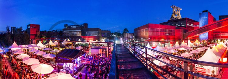 Einmal im Jahr treffen die regionalen Spitzengastronomen zur Gourmet-Meile Metropole Ruhr auf dem ehemaligen Gleisboulevard auf dem UNESCO-Welterbe Zollverein zusammen. Neben einer breiten Vielfalt kulinarischer Verlockungen bietet dieses Event ein anregendes und stimmungsvolles Ambiente für Gäste und Feinschmecker aus der ganzen Region.

Gleisboulevard nennt sich der Platz nördlich der Kohlenwäsche [A14] zwischen der Fritz-Schupp-Allee und der Halle 12 [A12]. Zu Betriebszeiten reihten sich dort die Waggons aneinander, die unter den Produktionsanlagen mit Kohle bzw. Bergematerial beladen wurden. 

Ort: Areal A [Schacht XII]

Das 55 Meter hohe Doppelbock-Fördergerüst ist das Wahrzeichen der Zeche Zollverein, der Stadt Essen und der gesamten Region: Die als Doppelstrebengerüst in Vollwandbauweise konstruierte Förderanlage ermöglichte eine doppelte Fördermechanik, mittels derer die doppelte Menge Förderwagen bzw. Fördergefäße transportiert werden konnten. Zollverein Schacht XII galt zu Betriebszeiten als die leistungsstärkste Zeche der Welt. 250 Stufen führen auf die oberste Plattform des Gerüstes, deren Seilscheiben sich auch heute noch drehen: Der Schacht wird heute von der RAG AG für die zentrale Wasserhaltung genutzt.

Pause machen im Bistro „Butterzeit!“ und Kunsthandwerk entdecken, wo früher Waggons mit Kohle beladen wurden: Die Ateliers im Erdgeschoss der Halle 12 [A12], darunter SchmuckProdukt und die ARKA Kulturwerkstatt, präsentieren neben eigenen Arbeiten auch wechselnde Ausstellungen anderer Künstler. Im Obergeschoss der ehemaligen Lesebandhalle finden heute Kongresse, Messen und Konzerte statt. Bis in die 1960er Jahre lasen hier Bergleute an fünf Lesebändern von Hand die großen Gesteinsstücke aus dem Rohkohlegemisch.

Der Wagenumlauf [A18] befindet sich zwischen der Wipperhalle und der Rückseite des Doppelbock-Fördergerüstes [A1]. In dem langgestreckten Gebäude wurden die Förderwagen, nachdem sie entleert wurden, wieder zurück zum Schacht geführt. Heute ist der Wagenumlauf Teil des Denkmalpfads ZOLLVEREIN®.

In den Kesselaschebunker [A13] (auch Aschebunker) wurde zu Betriebszeiten der Zeche über eine Rohrleitung aus dem Kesselhaus Asche eingeblasen. Die Kesselasche konnte vom Aschebunker direkt auf Eisenbahnwaggons verladen werden. Seit 2002 ist die begehbare Skulptur LA PRIMAVERA der deutsch-amerikanischen Künstlerin Maria Nordman in die ehemalige Durchfahrt des Bunkers installiert.

Zu Betriebszeiten der Zeche diente die Halle 10 [A10] als Lagerhaus und mechanische Werkstatt. Es befanden sich dort auch Meisterbüros. Heute sind hier Büros sowie das standorteigene Facility Management untergebracht.

Das Red Dot Design Museum präsentiert im ehemaligen Kesselhaus [A7] die weltweit größte Ausstellung zeitgenössischen Designs. Mehr als 2.000 Exponate, die alle mit dem Red Dot Design Award prämiert wurden, sind auf fünf Etagen zu bestaunen. Einst wurde hier Dampf erzeugt, der zum Antrieb von Druckluftkompressoren benötigt wurde. Der britische Architekt Norman Foster hat das Kesselhaus von 1992 bis 1996 umgebaut.

In der heutigen Ausstellungshalle [A8] produzierten früher Kompressoren Hochdruck, der zum Betrieb von Loks und anderen Großgeräten unter Tage verwendet wurde.