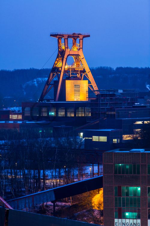 Die Geschichte Zollvereins beginnt in den Pioniertagen des heutigen Ruhrgebiets. Der erste Schacht wurde 1847 abgeteuft, d.h. in die Erde getrieben. Vier Jahre später wurde die erste Kohle gefördert. Im Laufe der Zeit entstanden vier weitere Schachtanlagen. Die letzte war die neue Zentralschachtanlage Zollverein XII, die ab 1932 die gesamte Zollverein-Kohle förderte. 

Noch heute besticht die symmetrische Anordnung der Gebäude auf zwei Blickachsen. Die komplett erhaltenen 20 Einzelgebäude bilden die technischen Arbeits- und Produktionsabläufe nach dem Prinzip „form follows function“ ab. Durchgestaltet bis in die Details der Lampen, Treppengeländer und Türgriffe, ist die Anlage ein komplett erhaltenes Gesamtkunstwerk. Die authentisch erhaltenen Anlagen repräsentieren exemplarisch die soziale, ökonomische, ästhetische und industrielle Geschichte des Kohle- und Stahlzeitalters. 

Trotz Rekordzahlen wie 12.000 Tagestonnen verwertbarer Steinkohle machte das Ende der Schwerindustrie auch vor Zollverein nicht Halt. Am 23. Dezember 1986 fuhr die letzte Schicht ein. An diesem Tag stand die Zeche bereits unter Denkmalschutz, 1990 begannen die Sanierungsarbeiten, seit 2001 sind Zeche und Kokerei Zollverein UNESCO-Welterbe. Mit dem Denkmalpfad ZOLLVEREIN®, dem Ruhr Museum und dem Red Dot Design Museum sind auf Schacht XII drei der beliebtesten Touristenziele der Region. Das Doppelbock-Fördergerüst der Schachtanlage ist heute Wahrzeichen für das gesamte Ruhrgebiet. 

Das 55 Meter hohe Doppelbock-Fördergerüst ist das Wahrzeichen der Zeche Zollverein, der Stadt Essen und der gesamten Region: Die als Doppelstrebengerüst in Vollwandbauweise konstruierte Förderanlage ermöglichte eine doppelte Fördermechanik. Dadurch konnte die doppelte Menge an Förderwagen bzw. Fördergefäßen transportiert werden. Zollverein Schacht XII galt zu Betriebszeiten als die leistungsstärkste Zeche der Welt. 250 Stufen führen auf die oberste Plattform des Gerüstes, deren Seilscheiben sich auch heute noch manchmal drehen: Der Schacht wird von der RAG AG für die zentrale Wasserhaltung genutzt.

Die diagonal und waagerecht zwischen den Gebäuden verlaufenden Bandbrücken sind fester Bestandteil der Gebäudeensembles von Zeche und Kokerei Zollverein. In ihrem Inneren befinden sich Bänder, auf denen die Rohkohle bzw. Kokskohle oder Koks zu den einzelnen Verarbeitungsstationen transportiert wurden. 

Areal A [Schacht XII]