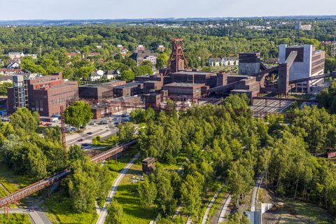 Die Geschichte Zollvereins beginnt in den Pioniertagen des heutigen Ruhrgebiets. Der erste Schacht wurde 1847 abgeteuft, d.h. in die Erde getrieben. Vier Jahre später wurde die erste Kohle gefördert. Im Laufe der Zeit entstanden vier weitere Schachtanlagen. Die letzte war die neue Zentralschachtanlage Zollverein XII, die ab 1932 die gesamte Zollverein-Kohle förderte. 

Noch heute besticht die symmetrische Anordnung der Gebäude auf zwei Blickachsen. Die komplett erhaltenen 20 Einzelgebäude bilden die technischen Arbeits- und Produktionsabläufe nach dem Prinzip „form follows function“ ab. Durchgestaltet bis in die Details der Lampen, Treppengeländer und Türgriffe, ist die Anlage ein komplett erhaltenes Gesamtkunstwerk. Die authentisch erhaltenen Anlagen repräsentieren exemplarisch die soziale, ökonomische, ästhetische und industrielle Geschichte des Kohle- und Stahlzeitalters. 

Trotz Rekordzahlen wie 12.000 Tagestonnen verwertbarer Steinkohle machte das Ende der Schwerindustrie auch vor Zollverein nicht Halt. Am 23. Dezember 1986 fuhr die letzte Schicht ein. An diesem Tag stand die Zeche bereits unter Denkmalschutz, 1990 begannen die Sanierungsarbeiten, seit 2001 sind Zeche und Kokerei Zollverein UNESCO-Welterbe. Mit dem Denkmalpfad ZOLLVEREIN®, dem Ruhr Museum und dem Red Dot Design Museum sind auf Schacht XII drei der beliebtesten Touristenziele der Region. Das Doppelbock-Fördergerüst der Schachtanlage ist heute Wahrzeichen für das gesamte Ruhrgebiet. 

Areal A [Schacht XII]

Das 55 Meter hohe Doppelbock-Fördergerüst ist das Wahrzeichen der Zeche Zollverein, der Stadt Essen und der gesamten Region: Die als Doppelstrebengerüst in Vollwandbauweise konstruierte Förderanlage ermöglichte eine doppelte Fördermechanik. Dadurch konnte die doppelte Menge an Förderwagen bzw. Fördergefäßen transportiert werden. Zollverein Schacht XII galt zu Betriebszeiten als die leistungsstärkste Zeche der Welt. 250 Stufen führen auf die oberste Plattform des Gerüstes, deren Seilscheiben sich auch heute noch manchmal drehen: Der Schacht wird von der RAG AG für die zentrale Wasserhaltung genutzt.

Areal A [Schacht XII], Fördergerüst [A1]

Das größte Übertagegebäude auf Zollverein, die Kohlenwäsche [A14], war zu Betriebszeiten eine Riesenmaschine: Täglich wurden dort 23.000 Tonnen Rohkohle sortiert und klassiert. Von 2003 bis 2006 wurde die Kohlenwäsche vom Office for Metropolitan Architecture (OMA) unter Leitung von Rem Koolhaas und dem Essener Büro Böll & Krabel umfassend saniert und für die spätere Nutzung vorbereitet. 

Eine 58 Meter lange Rolltreppe bringt die Besucher heute direkt in das RUHR.VISITORCENTER Essen auf der 24-Meter-Ebene. 2010 eröffnete das Ruhr Museum in der Kohlenwäsche, das in drei Etagen eine Dauerausstellung zu den Kategorien Gegenwart – Gedächtnis – Geschichte präsentiert. Die oberen Etagen gehören mit den authentisch erhaltenen Übertageanlagen zum Denkmalpfad ZOLLVEREIN®. Auf dem Dach der Kohlenwäsche sind zwei Aussichtsplattformen installiert, die einen Rundblick auf das Gelände und die Region bieten. Das Portal der Industriekultur informiert über industriekulturelle Angebote im Ruhrgebiet und in Nordrhein-Westfalen.

Areal A [Schacht XII], Kohlenwäsche [A14]