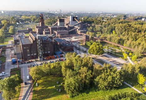 Die Geschichte Zollvereins beginnt in den Pioniertagen des heutigen Ruhrgebiets. Der erste Schacht wurde 1847 abgeteuft, d.h. in die Erde getrieben. Vier Jahre später wurde die erste Kohle gefördert. Im Laufe der Zeit entstanden vier weitere Schachtanlagen. Die letzte war die neue Zentralschachtanlage Zollverein XII, die ab 1932 die gesamte Zollverein-Kohle förderte. 

Noch heute besticht die symmetrische Anordnung der Gebäude auf zwei Blickachsen. Die komplett erhaltenen 20 Einzelgebäude bilden die technischen Arbeits- und Produktionsabläufe nach dem Prinzip „form follows function“ ab. Durchgestaltet bis in die Details der Lampen, Treppengeländer und Türgriffe, ist die Anlage ein komplett erhaltenes Gesamtkunstwerk. Die authentisch erhaltenen Anlagen repräsentieren exemplarisch die soziale, ökonomische, ästhetische und industrielle Geschichte des Kohle- und Stahlzeitalters. 

Trotz Rekordzahlen wie 12.000 Tagestonnen verwertbarer Steinkohle machte das Ende der Schwerindustrie auch vor Zollverein nicht Halt. Am 23. Dezember 1986 fuhr die letzte Schicht ein. An diesem Tag stand die Zeche bereits unter Denkmalschutz, 1990 begannen die Sanierungsarbeiten, seit 2001 sind Zeche und Kokerei Zollverein UNESCO-Welterbe. Mit dem Denkmalpfad ZOLLVEREIN®, dem Ruhr Museum und dem Red Dot Design Museum sind auf Schacht XII drei der beliebtesten Touristenziele der Region. Das Doppelbock-Fördergerüst der Schachtanlage ist heute Wahrzeichen für das gesamte Ruhrgebiet. 

Areal A [Schacht XII]