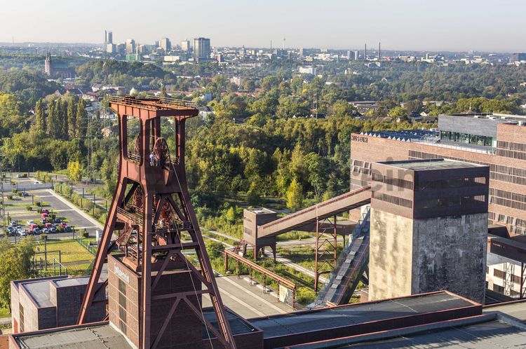 Die Geschichte Zollvereins beginnt in den Pioniertagen des heutigen Ruhrgebiets. Der erste Schacht wurde 1847 abgeteuft, d.h. in die Erde getrieben. Vier Jahre später wurde die erste Kohle gefördert. Im Laufe der Zeit entstanden vier weitere Schachtanlagen. Die letzte war die neue Zentralschachtanlage Zollverein XII, die ab 1932 die gesamte Zollverein-Kohle förderte. 

Noch heute besticht die symmetrische Anordnung der Gebäude auf zwei Blickachsen. Die komplett erhaltenen 20 Einzelgebäude bilden die technischen Arbeits- und Produktionsabläufe nach dem Prinzip „form follows function“ ab. Durchgestaltet bis in die Details der Lampen, Treppengeländer und Türgriffe, ist die Anlage ein komplett erhaltenes Gesamtkunstwerk. Die authentisch erhaltenen Anlagen repräsentieren exemplarisch die soziale, ökonomische, ästhetische und industrielle Geschichte des Kohle- und Stahlzeitalters. 

Trotz Rekordzahlen wie 12.000 Tagestonnen verwertbarer Steinkohle machte das Ende der Schwerindustrie auch vor Zollverein nicht Halt. Am 23. Dezember 1986 fuhr die letzte Schicht ein. An diesem Tag stand die Zeche bereits unter Denkmalschutz, 1990 begannen die Sanierungsarbeiten, seit 2001 sind Zeche und Kokerei Zollverein UNESCO-Welterbe. Mit dem Denkmalpfad ZOLLVEREIN®, dem Ruhr Museum und dem Red Dot Design Museum sind auf Schacht XII drei der beliebtesten Touristenziele der Region. Das Doppelbock-Fördergerüst der Schachtanlage ist heute Wahrzeichen für das gesamte Ruhrgebiet. 

Das 55 Meter hohe Doppelbock-Fördergerüst ist das Wahrzeichen der Zeche Zollverein, der Stadt Essen und der gesamten Region: Die als Doppelstrebengerüst in Vollwandbauweise konstruierte Förderanlage ermöglichte eine doppelte Fördermechanik. Dadurch konnte die doppelte Menge an Förderwagen bzw. Fördergefäßen transportiert werden. Zollverein Schacht XII galt zu Betriebszeiten als die leistungsstärkste Zeche der Welt. 250 Stufen führen auf die oberste Plattform des Gerüstes, deren Seilscheiben sich auch heute noch manchmal drehen: Der Schacht wird von der RAG AG für die zentrale Wasserhaltung genutzt.

Das größte Übertagegebäude auf Zollverein, die Kohlenwäsche [A14], war zu Betriebszeiten eine Riesenmaschine: Täglich wurden dort 23.000 Tonnen Rohkohle sortiert und klassiert. Von 2003 bis 2006 wurde die Kohlenwäsche vom Office for Metropolitan Architecture (OMA) unter Leitung von Rem Koolhaas und dem Essener Büro Böll & Krabel umfassend saniert und für die spätere Nutzung vorbereitet. 

Eine 58 Meter lange Rolltreppe bringt die Besucher heute direkt in das RUHR.VISITORCENTER Essen auf der 24-Meter-Ebene. 2010 eröffnete das Ruhr Museum in der Kohlenwäsche, das in drei Etagen eine Dauerausstellung zu den Kategorien Gegenwart – Gedächtnis – Geschichte präsentiert. Die oberen Etagen gehören mit den authentisch erhaltenen Übertageanlagen zum Denkmalpfad ZOLLVEREIN®. Auf dem Dach der Kohlenwäsche sind zwei Aussichtsplattformen installiert, die einen Rundblick auf das Gelände und die Region bieten. Das Portal der Industriekultur informiert über industriekulturelle Angebote im Ruhrgebiet und in Nordrhein-Westfalen.

Areal A [Schacht XII]