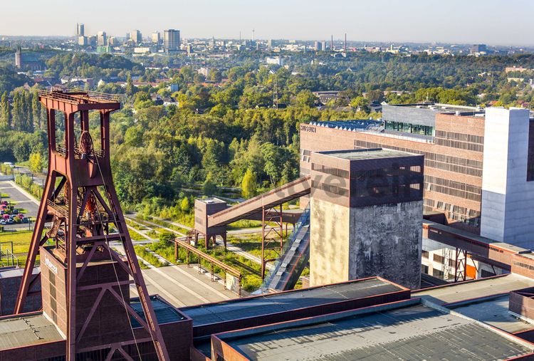 Die Geschichte Zollvereins beginnt in den Pioniertagen des heutigen Ruhrgebiets. Der erste Schacht wurde 1847 abgeteuft, d.h. in die Erde getrieben. Vier Jahre später wurde die erste Kohle gefördert. Im Laufe der Zeit entstanden vier weitere Schachtanlagen. Die letzte war die neue Zentralschachtanlage Zollverein XII, die ab 1932 die gesamte Zollverein-Kohle förderte. 

Noch heute besticht die symmetrische Anordnung der Gebäude auf zwei Blickachsen. Die komplett erhaltenen 20 Einzelgebäude bilden die technischen Arbeits- und Produktionsabläufe nach dem Prinzip „form follows function“ ab. Durchgestaltet bis in die Details der Lampen, Treppengeländer und Türgriffe, ist die Anlage ein komplett erhaltenes Gesamtkunstwerk. Die authentisch erhaltenen Anlagen repräsentieren exemplarisch die soziale, ökonomische, ästhetische und industrielle Geschichte des Kohle- und Stahlzeitalters. 

Trotz Rekordzahlen wie 12.000 Tagestonnen verwertbarer Steinkohle machte das Ende der Schwerindustrie auch vor Zollverein nicht Halt. Am 23. Dezember 1986 fuhr die letzte Schicht ein. An diesem Tag stand die Zeche bereits unter Denkmalschutz, 1990 begannen die Sanierungsarbeiten, seit 2001 sind Zeche und Kokerei Zollverein UNESCO-Welterbe. Mit dem Denkmalpfad ZOLLVEREIN®, dem Ruhr Museum und dem Red Dot Design Museum sind auf Schacht XII drei der beliebtesten Touristenziele der Region. Das Doppelbock-Fördergerüst der Schachtanlage ist heute Wahrzeichen für das gesamte Ruhrgebiet. 

Das 55 Meter hohe Doppelbock-Fördergerüst ist das Wahrzeichen der Zeche Zollverein, der Stadt Essen und der gesamten Region: Die als Doppelstrebengerüst in Vollwandbauweise konstruierte Förderanlage ermöglichte eine doppelte Fördermechanik. Dadurch konnte die doppelte Menge an Förderwagen bzw. Fördergefäßen transportiert werden. Zollverein Schacht XII galt zu Betriebszeiten als die leistungsstärkste Zeche der Welt. 250 Stufen führen auf die oberste Plattform des Gerüstes, deren Seilscheiben sich auch heute noch manchmal drehen: Der Schacht wird von der RAG AG für die zentrale Wasserhaltung genutzt.

Das größte Übertagegebäude auf Zollverein, die Kohlenwäsche [A14], war zu Betriebszeiten eine Riesenmaschine: Täglich wurden dort 23.000 Tonnen Rohkohle sortiert und klassiert. Von 2003 bis 2006 wurde die Kohlenwäsche vom Office for Metropolitan Architecture (OMA) unter Leitung von Rem Koolhaas und dem Essener Büro Böll & Krabel umfassend saniert und für die spätere Nutzung vorbereitet. 

Eine 58 Meter lange Rolltreppe bringt die Besucher heute direkt in das RUHR.VISITORCENTER Essen auf der 24-Meter-Ebene. 2010 eröffnete das Ruhr Museum in der Kohlenwäsche, das in drei Etagen eine Dauerausstellung zu den Kategorien Gegenwart – Gedächtnis – Geschichte präsentiert. Die oberen Etagen gehören mit den authentisch erhaltenen Übertageanlagen zum Denkmalpfad ZOLLVEREIN®. Auf dem Dach der Kohlenwäsche sind zwei Aussichtsplattformen installiert, die einen Rundblick auf das Gelände und die Region bieten. Das Portal der Industriekultur informiert über industriekulturelle Angebote im Ruhrgebiet und in Nordrhein-Westfalen.

Areal A [Schacht XII]